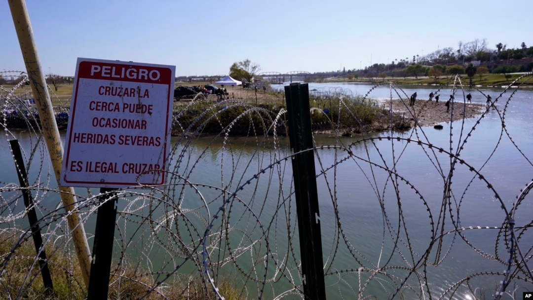 Migrants cross the Rio Grande into the US from Mexico behind concertina wire and a sign warning that it's dangerous and illegal to cross, Jan. 3, 2024, in Eagle Pass, Texas. 