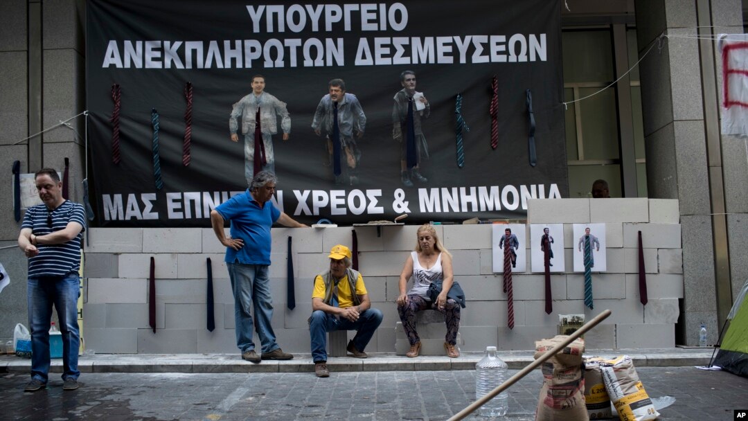 Protesting hospital staff sit in front of a wall that they built at the entrance of the Greek Finance Ministry with a banner depicting Greek Prime Minister Alexis Thipras, Deputy Health Minister Pavlos Polakis and Greek Finance Minister Euclid Tsakalotos wearing ties reading ''Ministry of broken promises" and " We drown in debt and bailouts" in central Athens, June 16, 2017.