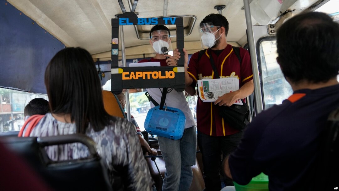 Juan Pablo Lares, right, holds a cardboard frame in front of his associate Maximiliano Bruzual who reads their newscast "El Bus TV Capitolio" to commuters on a bus in Caracas, Venezuela. (AP Photo/Ariana Cubillos)